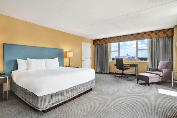 Photo of luxury modern hotel room with king size bed, tv in front of it and big window with curtains and a desk set up and sofa chair in front of the window