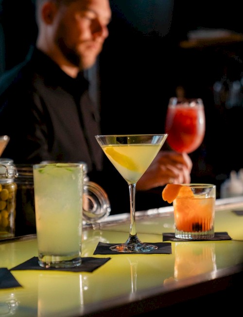 photo of bar counter with multiple colorful cocktails and bartender handing a glass of a red cocktail