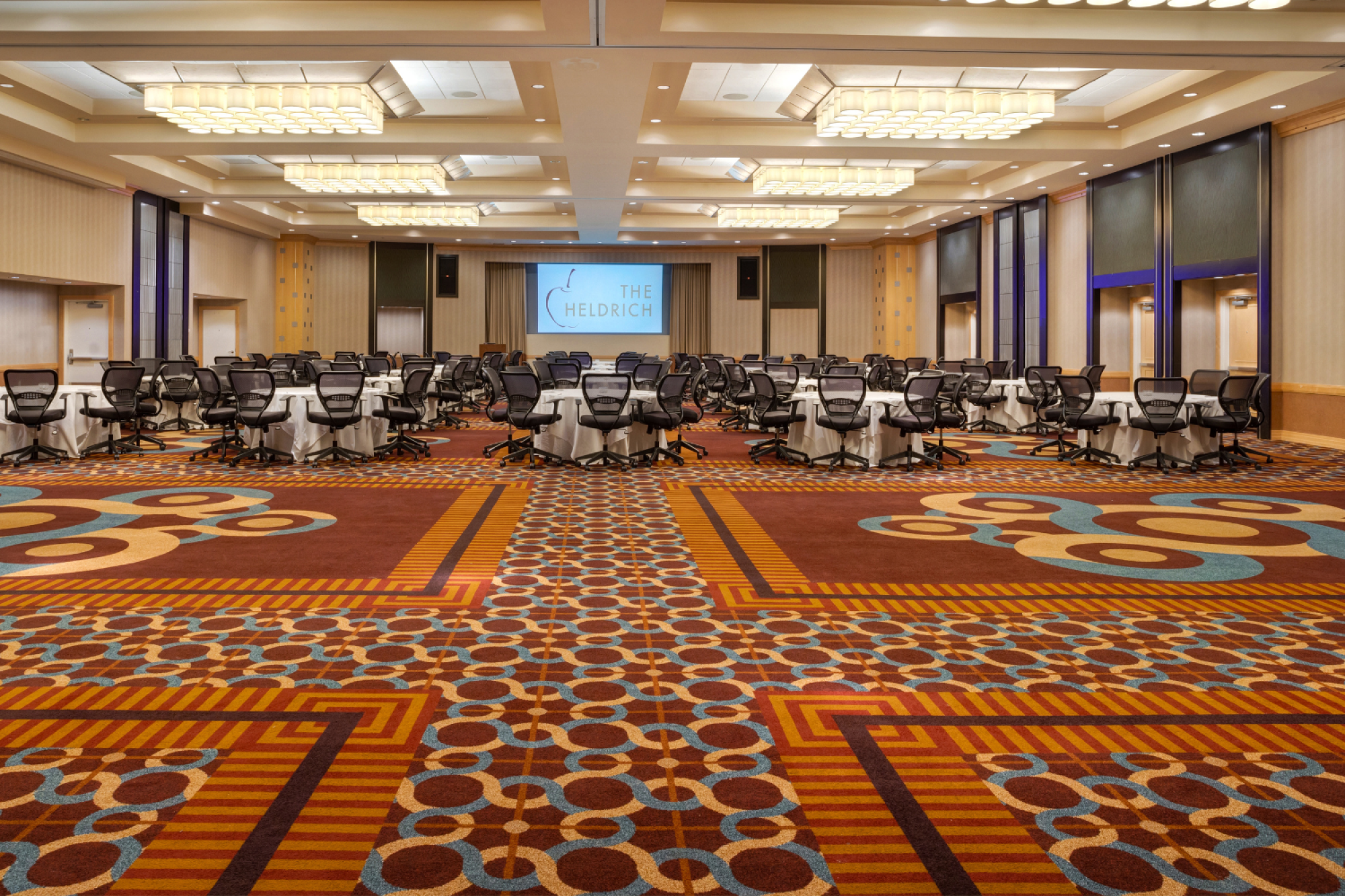 Photo of empty carpeted ballroom with multiple ceiling lights and a few chairs and tables in the back of the room