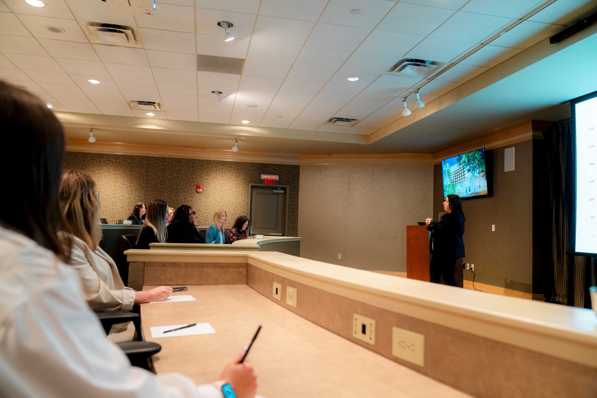 A person giving a presentation in a conference room, with attendees seated and taking notes, while a screen displays content.