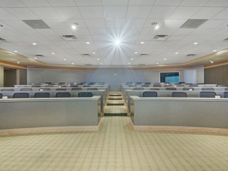 Photo of meeting room with multiple lines of long tables and chairs in a light wooden floor with a ceiling projector