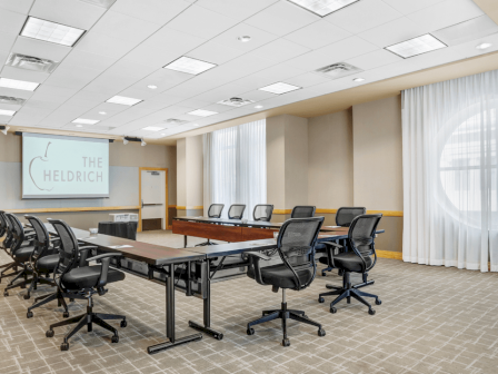 Photo of meeting room in u set up with black wheeled chairs, multiple windows and a white screen to project in the back of the meeting room