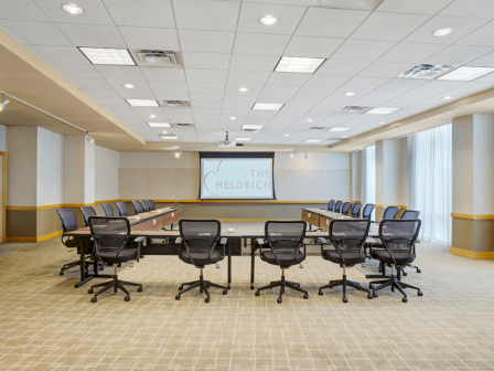 Photo of meeting room in u set up with black wheeled chairs, multiple windows and a white screen to project in the back of the meeting room