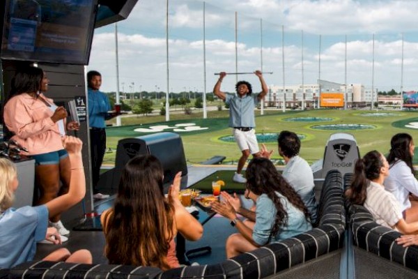 Photo of some people playing top golf while others are sitting on some sofas and raising their hands celebrating