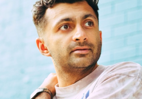 A person with short hair and a watch looks to the side, against a blue brick background, wearing a casual light-colored shirt.