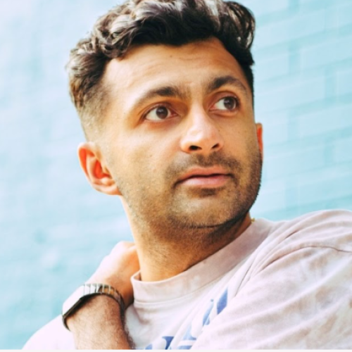 A person with short hair and a watch looks to the side, against a blue brick background, wearing a casual light-colored shirt.