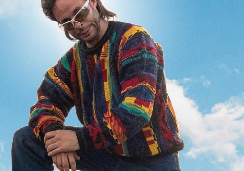 A person in a colorful sweater and sunglasses poses against a blue sky with clouds.