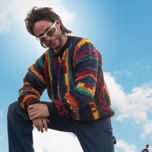 A person in a colorful sweater and sunglasses poses against a blue sky with clouds.