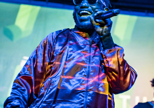 A person wearing sunglasses and a shiny jacket performs on stage, holding a microphone, with a vibrant background.