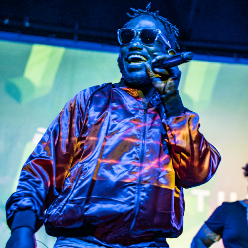 A person wearing sunglasses and a shiny jacket performs on stage, holding a microphone, with a vibrant background.