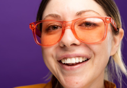 A person wearing orange glasses and a yellow jacket smiles against a purple background.