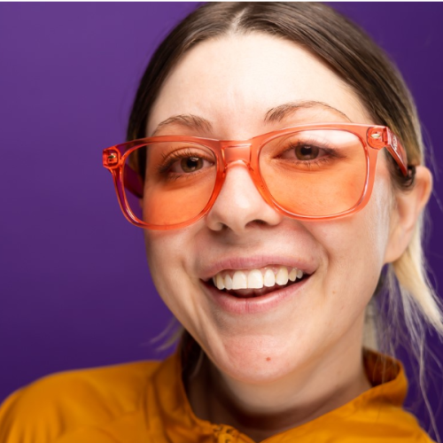 A person wearing orange glasses and a yellow jacket smiles against a purple background.