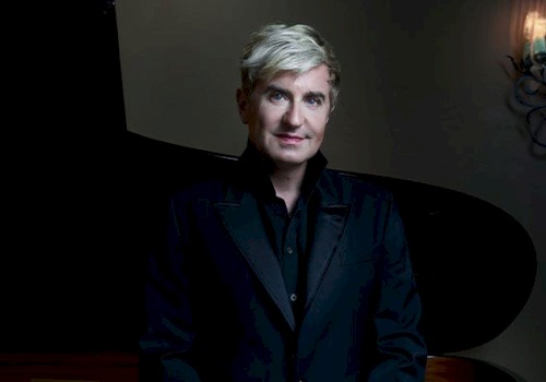 A person with short blonde hair wearing a black suit stands in front of a grand piano, with a softly lit background.