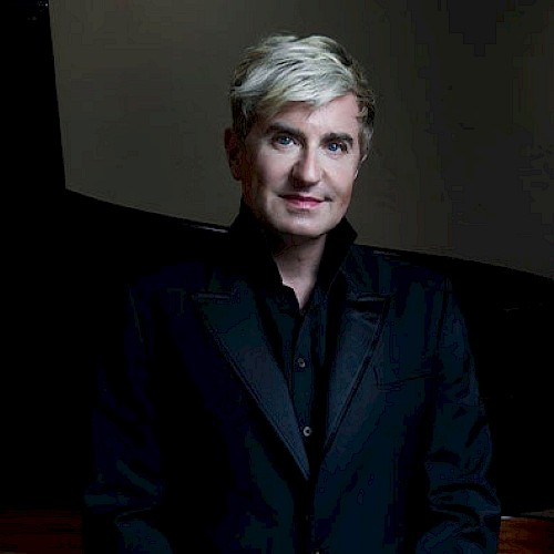 A person with short blonde hair wearing a black suit stands in front of a grand piano, with a softly lit background.