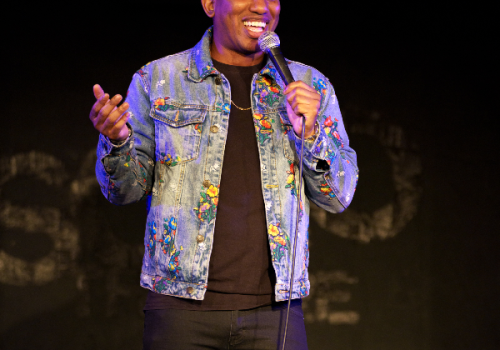 A person on stage holding a microphone, wearing a red cap and a blue patterned denim jacket, is smiling and talking.