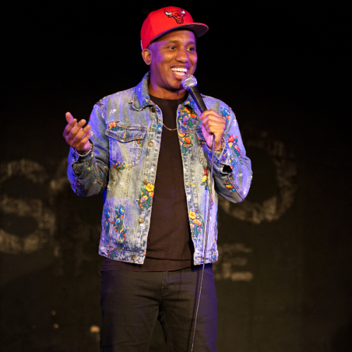 A person on stage holding a microphone, wearing a red cap and a blue patterned denim jacket, is smiling and talking.