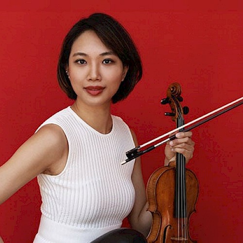 A person in a white dress holding a violin against a bright red background.