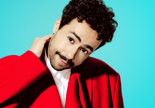 A person with curly hair and a mustache is wearing a red coat, posing against a light blue background.