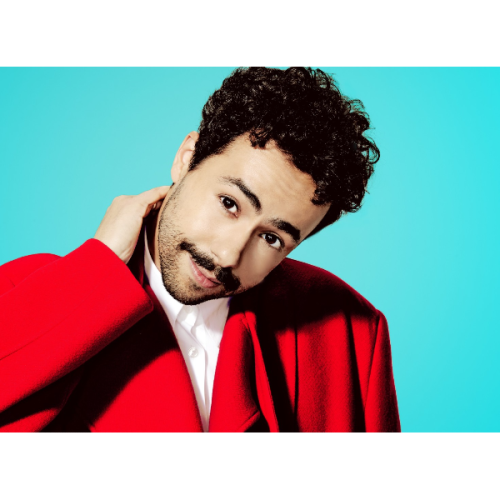 A person with curly hair and a mustache is wearing a red coat, posing against a light blue background.