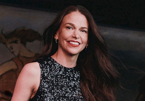 A person with long brown hair and a black sequined top smiling, with an artistic background.