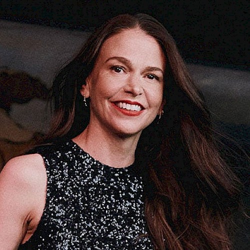 A person with long brown hair and a black sequined top smiling, with an artistic background.