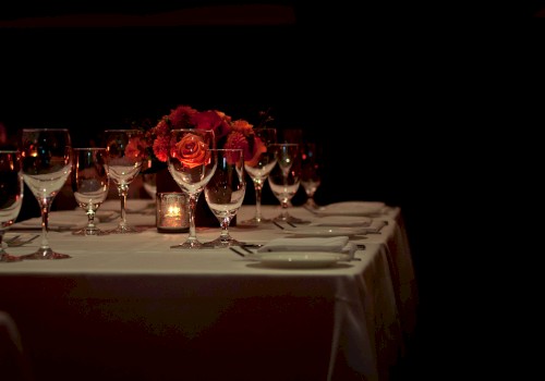 A dimly lit, elegantly set dining table with roses, glasses, and a candle centerpiece, ready for a formal gathering or dinner event.