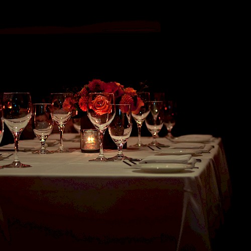 A dimly lit, elegantly set dining table with roses, glasses, and a candle centerpiece, ready for a formal gathering or dinner event.