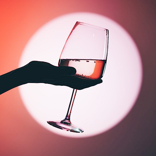 A hand holding a wine glass with a pink beverage, silhouetted against a large, out-of-focus circular light in the background.