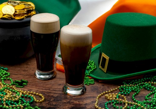 Two pints of beer, a green hat, beads, and a cauldron of gold coins are set against an Irish flag backdrop, suggesting a festive atmosphere.