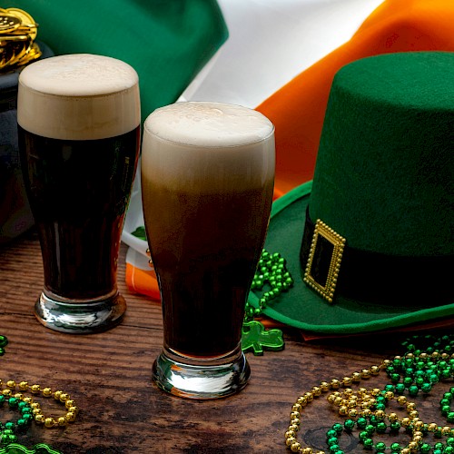 Two pints of beer, a green hat, beads, and a cauldron of gold coins are set against an Irish flag backdrop, suggesting a festive atmosphere.