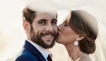 A bride kisses a smiling groom under a veil, capturing a tender moment.