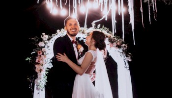 A couple smiling and embracing in front of a floral arch, with hanging ribbons and lights, during a wedding ceremony.